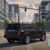 "A realistic 3D driving simulation game screen set in an urban environment. The camera is positioned behind a black SUV, showing a rear and side view of the vehicle as it waits at a traffic light. The scene features tall modern buildings with glass windows, balconies, and detailed facades. In the background, a clear sky with light clouds is visible, along with traffic lights hanging from poles. The traffic light shows a yellow signal, while a pedestrian light below shows a red 'stop' symbol. On the left side of the screen, a circular speedometer gauge is displayed, with a light blue border. The needle points to 1, and the speed is indicated as '0 KM/H.' A large, semi-transparent steering wheel icon is placed at the bottom-left corner, representing the control input for steering. On the right side, two semi-transparent icons represent car pedals: one for acceleration (depicted by a gas pedal icon) and the other for braking (depicted by a brake pedal icon). The environment includes trees, fences, and shadows on the pavement, adding to the realistic urban atmosphere. The buildings in the distance have modern architectural designs, with some featuring unique curved blue accents."